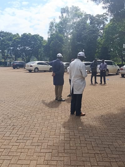 The image depicts an outdoor scene in Kaplelwo, Kenya, on a bright, sunny day with a partly cloudy sky. The ground is covered with light brown, rectangular brick pavers laid in a herringbone pattern.
In the foreground, facing away from the viewer, are two men. The man on the left wears a dark, long-sleeved jacket or coat, light-colored trousers, and a white baseball cap. His right arm is slightly raised, and he appears to be pointing towards the left side of the frame. To his right, another man wears a light-colored, possibly white, long-sleeved shirt or lab coat, dark trousers, and a white baseball cap.
Further in the background, a small group of three to four men stands facing generally towards the right. They are dressed in casual attire, including dark shirts and trousers.
Behind the groups of people, several vehicles are parked. These include a white sedan, a dark SUV, and a light-colored van. The background is dominated by numerous large, green trees, suggesting a lush, vegetated environment. Shadows of the people and possibly some trees are cast on the paved ground, indicating the sun's position. No visible text is present in the image. The scene suggests a casual gathering or discussion taking place outdoors in what appears to be a parking lot or open communal area.