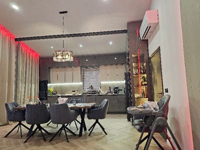 This image captures a modern, open-plan dining and kitchen area within a residence in the city of Dragushinovo, Bulgaria. The central focus is a rectangular wooden dining table with a black metal base, surrounded by six gray upholstered chairs featuring black star-shaped leg structures. The table is set with placemats and includes a small vase holding orange flowers. Overhead, a multi-tiered circular chandelier with crystal or glass elements hangs, complemented by recessed ceiling lights. Along the left wall, cream-colored curtains are accented with red LED strip lighting running along the top.
The kitchen area, visible in the background, features gray base cabinets, white upper cabinets, and a white tiled backsplash illuminated by under-cabinet lighting. A black coffee machine and other small appliances are present on the countertop. To the right, a white wall-mounted air conditioning unit is positioned above a tall, narrow display cabinet with glass shelving, housing various bottles and illuminated by vertical red LED strips. Adjacent to this cabinet is a dark brown door with a horizontal metallic accent and a black handle. In the immediate right foreground, a gray and red baby high chair is visible, equipped with a tray holding unspecified items covered by a white cloth. The flooring throughout the space is a light-colored wood or laminate with a herringbone pattern. No individuals are present, and no actions or interactions are depicted.