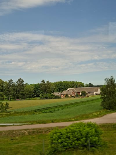 The image captures a rural landscape under a partly cloudy blue sky. In the distance, a farm complex with several connected buildings is visible, featuring a red-brick section and larger, gray-roofed structures. A red tractor is parked near one of the buildings. The farm is surrounded by lush green fields, some with tilled rows, and bordered by a dense line of trees. In the foreground, a paved road curves through the scene, with a grassy verge and some bushes along its edge. The overall impression is one of a peaceful, working farmstead in a scenic countryside setting. The provided context suggests this location is in Lipnik, Poland.
