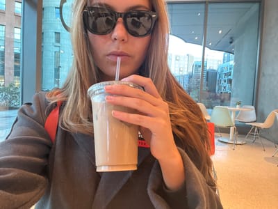 A person wearing sunglasses holds a tall plastic cup with a straw. The cup appears to contain a light brown beverage. The individual wears a dark gray jacket and an orange lanyard around their neck. A large window reveals a city skyline with buildings in the background. A light fixture with multiple tiers of bowls is visible on the ceiling. The picture is rotated.