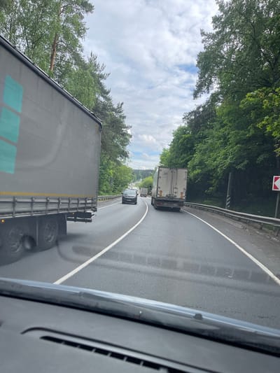 The image shows a view from inside a vehicle, likely a truck, traveling on a wet, winding road. In the foreground, a large truck is visible, taking up much of the lower half of the frame. Ahead of the truck, a black car is following another truck. The road curves to the right and is bordered by a metal guardrail. Beyond the guardrail, dense green trees line the road. A road sign is visible on the right side of the road. The sky is overcast with patches of blue visible through the trees. It is not possible to confirm the location within Semyonovskoye, Russia without additional information.