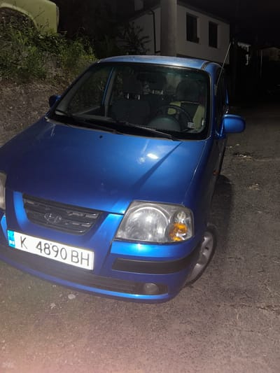 A blue car, with a Bulgarian license plate that reads "K 4890 BH," is parked on the side of what appears to be a paved road. The car is facing towards the viewer's right. A building is visible in the background on the left side of the image. The setting is nighttime. The location might be in the city of Pripek, Bulgaria.