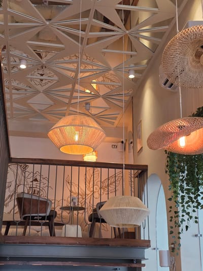 The image captures an interior scene, possibly a cafe or restaurant, in Tijuana, Mexico. The camera angle is tilted, presenting an unconventional perspective.

The scene is illuminated by several hanging light fixtures, crafted from materials like wicker and rope, and emitting a warm, orange glow. Some of the lamps are round and flat, while others are elongated and hexagonal.

A geometric, decorative screen with triangular cutouts is visible on one side, while the opposite side features a metal railing and a wall mural depicting coffee beans and other design elements. Seating is visible behind the railing, suggesting a dining area.

Green foliage is draped across the ceiling in the upper part of the frame, adding a touch of nature to the scene. Other ceiling-mounted fixtures and structural elements can be seen. The overall ambiance is warm and inviting.