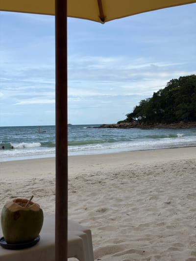 This image depicts a serene beach scene at Ao Cho, Thailand. The foreground is partially occupied by the edge of a yellow umbrella and a dark wooden pole, suggesting a shaded seating area. On the right, a partially opened green coconut with a straw rests on a white surface, likely a table or chair.
The middle ground reveals a sandy beach leading to the ocean. The sand exhibits ripples, indicating a gentle tide or wind. The ocean is a vibrant turquoise, with white waves lapping at the shore. Further out, a person on a paddleboard can be seen in the distance, and two other figures are in the water closer to the shore, suggesting recreational activities.
The background features a rocky shoreline covered in lush green trees, creating a dramatic contrast with the sandy beach and blue ocean. The sky is a pale blue with scattered white clouds, hinting at pleasant weather. The lighting suggests it's likely late afternoon or early morning. The overall mood is one of relaxation and tranquility, characteristic of a tropical beach destination. No text is visible in the image.
