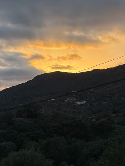 The image shows a mountainous landscape with a sunset sky. The sky is filled with dramatic clouds, with the top half being dark grey and the bottom half illuminated by the warm orange glow of the setting sun. A dark, tree-covered mountain slopes from the foreground into the background. Several houses are visible scattered on the mountainside, some appearing white. Two dark wires stretch horizontally across the frame, obscuring parts of the mountain and sky. The foreground is dominated by dark green foliage. The scene is located in the region of Loulé, Portugal.