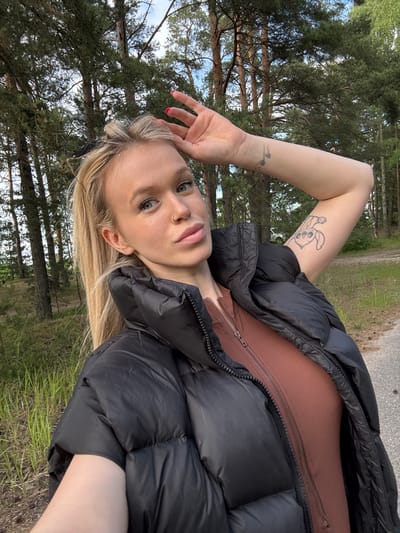 A young woman with blonde hair and blue eyes is taking a selfie outdoors. She is wearing a black puffer jacket over a brown form-fitting outfit. Her right arm is raised, with her hand resting on her head. A tattoo of a cartoon character is visible on her upper left arm. She is wearing a nose ring and has red nail polish on her fingers. The background consists of a forest with tall pine trees, green grass, and a paved path. The lighting suggests it is daytime, possibly late afternoon given the quality of light. The location appears to be the outskirts of Bolshaya Izhora, Russia, characterized by its coastal pine forests.