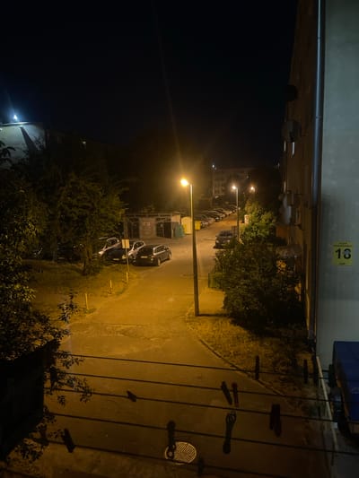 This is a nighttime photograph taken in Konstantynów Łódzki, Poland. The scene depicts a courtyard area between apartment buildings, illuminated by streetlights. In the foreground, clotheslines with clothespins are visible. Further into the scene, a paved path winds through a grassy area with trees. Several parked cars are present, and a low-rise building with windows is visible in the background. On the right side of the image, a section of a building facade is visible, featuring a vertical drainpipe and a yellow street sign displaying the number "18" and text. The lighting creates strong shadows, characteristic of a nighttime urban environment. No people are visible in the frame.