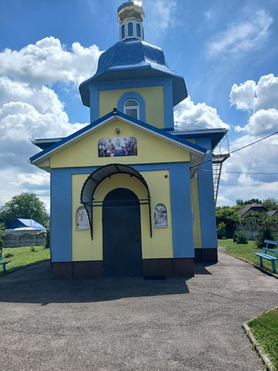 The image shows a small, brightly colored Ukrainian Orthodox church with blue and yellow walls and a blue, domed roof topped with a golden cupola. The church is situated in a semi-rural area. In front of the church, there is a paved walkway leading to a dark, arched doorway. Above the doorway is a religious mural depicting several figures. Smaller religious images are placed on either side of the doorway. To the left of the church, a chain-link fence with a concrete base separates it from a grassy area with a blue-covered structure in the distance. To the right, there is a grassy area with a blue bench. The sky is bright blue with fluffy white clouds. The scene appears to be located in the village of Doslidnytske, Ukraine, based on the provided context.