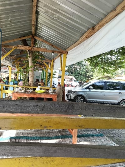 This image depicts a semi-open-air dining area, likely a restaurant or warung, situated in Gembong, Indonesia, during daylight hours. The structure features a corrugated metal roof supported by a frame of wooden beams and posts, some painted yellow. White canvas fabric is draped along the right side of the roof structure.
In the mid-ground, multiple individuals are present. Several people are seated on mats around low wooden tables, with food items, cups, and a yellow bowl visible on the tables. One prominent low wooden table in the center-left shows multiple plates and cups with dark liquid. To the right of this table, the lower legs and feet of a person, covered in light-colored fabric, are visible, indicating they are seated on a mat directly on the ground. A tree trunk with some foliage is integrated into the dining area's structure.
In the background, beyond a yellow railing, a silver multi-purpose vehicle (MPV) or small SUV is parked, facing left. Further behind it, a white commercial van, possibly a food service vehicle, and at least one motorcycle are visible. Green trees and foliage are present beyond the vehicles, indicating an outdoor setting. The foreground shows worn wooden planks, some painted yellow, forming a barrier or part of the seating arrangement, with woven mats visible on the ground beneath. A sign with partially visible Indonesian text, including "IKAN BAKAR" (grilled fish), is visible in the background on the left, further indicating a food establishment.