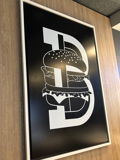 A vertical, framed artwork hangs on a light brown, wood-paneled wall. The artwork is a black rectangle with a white stylized graphic of a hamburger. The hamburger is depicted in profile, with its various layers, including the bun, patty, lettuce, and tomato, outlined in white. The design of the hamburger is also integrated with large, stylized white block letters, forming what appears to be the letter "B" or possibly an "8" with the hamburger components layered within it. The frame is white. The wall has a visible wood grain texture. The ceiling above the artwork is a textured gray acoustic tile.