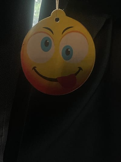 The image shows a close-up of a yellow smiley face air freshener hanging from a string. The air freshener has large blue eyes with white pupils and a wide mouth with its red tongue sticking out. The smiley face also has rosy cheeks. The background is dark and appears to be fabric, possibly from a car's interior. There is a vertical light source visible in the upper left corner of the image. There is no information in the image to indicate its location in Ganderkesee, Germany.