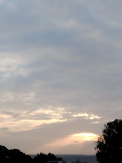 The scene depicts an outdoor landscape observed from Pyli, Greece, at either sunrise or sunset. The upper two-thirds of the image consist of a cloudy sky, featuring layered altocumulus-type formations ranging in color from light grey to muted blue-grey. Towards the lower right quadrant, the sun is partially obscured by clouds, emitting a concentrated burst of golden-orange light and visible rays extending horizontally and upwards through cloud breaks. A dark, indistinct horizontal line crosses the lower mid-section of the frame, representing a distant body of water or horizon line. Above this line, faint, dark, and blurred shapes suggest distant landmasses or structures. The immediate foreground, along the very bottom edge of the image, is occupied by dark, amorphous silhouettes of dense tree foliage on both the left and right sides. No individuals or specific human activities are observable.