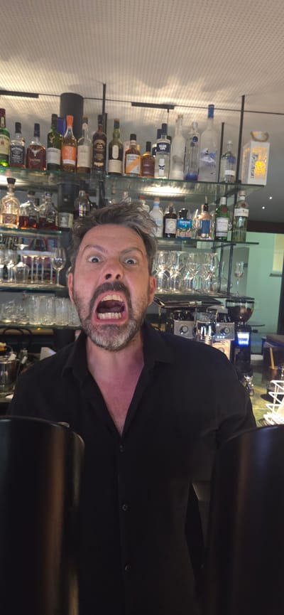 This media file shows a man with a surprised or shocked expression, his mouth wide open. He appears to be in a bar or restaurant setting, with shelves filled with bottles of alcohol behind him. The bottles are of various shapes and sizes, suggesting a wide selection of beverages. The man is wearing a black shirt. The lighting appears to be artificial, consistent with an indoor establishment. The image is oriented vertically, with the man's head towards the bottom. The scene suggests a moment of unexpected excitement or perhaps a joke being played. The man's grey hair and beard, along with his facial expression, are prominent details. The backdrop of shelves filled with bottles provides context for the location as a place that serves drinks.