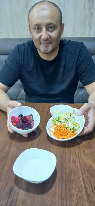 The image shows a man wearing a black t-shirt, sitting at a wooden table. He is holding two small white bowls. One bowl contains a salad of shredded carrots and cabbage. The other bowl holds diced beets and what appear to be radishes. To the right of these bowls is a third empty white bowl. The man's expression is neutral, and he appears to be indoors. The scene suggests a meal or a preparation for a meal. The background includes a grey sofa and a light-colored wall with a wood-grain pattern. The lighting suggests daytime. There is no visible text in the image. The location context is Khodja Akhrar Vali, Uzbekistan.
