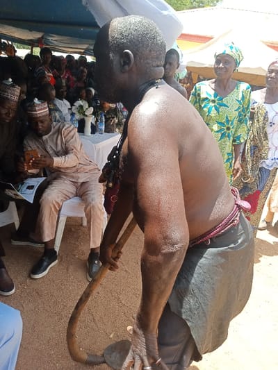 The image shows a man standing in a courtyard in Zum, Nigeria, holding a long wooden implement in his hands. He is wearing a black skirt and a red and white belt with straps over his shoulders. The man's back is to the camera. The man is looking to his left, where several people are sitting on white chairs under a blue canopy. The group of people to the left of the man is mostly men, wearing traditional clothing, and one woman in a hijab. Behind the man, to the right of the image, there are other women standing, wearing colorful clothing and headscarves. There is a large blue canopy behind the woman, and there appears to be some type of building in the background. The ground is dusty and dry. The image is likely taken during a public ceremony.
