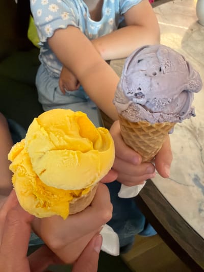 This image displays an indoor scene featuring two individuals holding ice cream cones. While the requested location is Dalaman, Turkey, no specific visual identifiers for this city are present in the image. In the immediate foreground, an adult hand holds a waffle cone containing a large scoop of bright yellow ice cream, with white napkin material partially visible beneath. Behind this, a child, wearing a light blue top with white daisy patterns and light blue lower garments, holds a separate waffle cone with a large scoop of light purple ice cream. The child's arms are positioned with one arm partially crossed over the other, holding the cone in the right hand. The environment includes a light-colored, possibly marble-patterned tabletop visible on the right, and a dark, likely upholstered, seating surface in the left background. The ice cream in both cones exhibits minor textural variations consistent with light melting.
