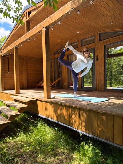 A young woman is captured mid-action, performing a dynamic dance leap or high kick on a spacious wooden deck. She is smiling, wearing sunglasses, a white long-sleeved top, dark blue leggings, and light-colored socks. Her left leg is extended forward and upward, while her right leg is bent backward, with her arms outstretched for balance.

The scene is set on a well-maintained wooden deck adjacent to a modern wooden house or cabin. The house features light-colored wood paneling and large dark-framed windows, some reflecting the green trees and blue sky. A light blue yoga mat is visible on the deck near the woman's right foot. Overhead, a string of spherical light bulbs decorates the wooden beams of the deck's ceiling.

Bright sunlight illuminates the scene, casting clear shadows and indicating a sunny daytime setting. Beyond the edge of the deck, lush green grass and foliage grow, suggesting a natural, possibly rural or secluded environment. Wooden stairs are partially visible, descending from the deck into the greenery. On the exterior of the house, a dark number "17" is visible near one of the window frames. The image conveys a sense of joy, energy, and outdoor activity. The location is contextualized as Vozhoiskii, Russia.