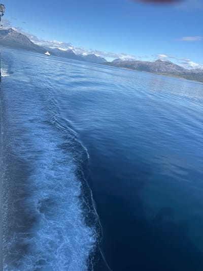 This media file captures a serene, high-angle view of a body of water, likely a fjord, with mountains lining the left side. The water is a deep blue, transitioning to a lighter, more rippled surface closer to the shore, where small waves are breaking. A white ship is visible in the distance, sailing on the calmer waters near the mountainous coastline. The sky above is a clear blue with a few scattered white clouds. The overall scene suggests a bright, sunny day, possibly in the late morning or early afternoon given the lighting. The presence of mountains and the clear blue water hints at a scenic, possibly Scandinavian, location, aligning with the context of Lodingen, Norway. No people are directly visible in the frame, and there is no visible text. The overall mood is peaceful and picturesque, emphasizing the natural beauty of the landscape.