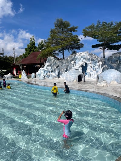 The scene depicts a water park in Kledung, Indonesia. The area includes a swimming pool with several people. One child, wearing a pink and light blue swimsuit and goggles, is immersed in the water. Another child, wearing a yellow shirt, stands in the shallow end. A third person, in dark clothing, is seen near a decorative rocky formation. The water park incorporates igloo-shaped structures along the pool's edge. The sky is blue with scattered clouds, and trees and a building with a red roof are visible in the background.