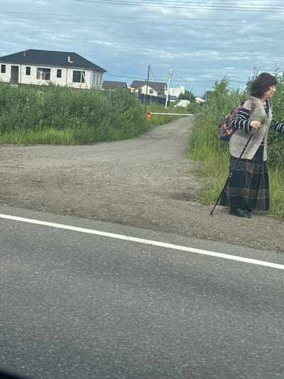 A woman stands on a gravel path next to a paved road with a white line. She is wearing a grey knit vest over a black and white striped long-sleeved shirt and a long, dark plaid skirt. She has a patterned backpack slung over her shoulder and is holding a black cane. Her head is tilted back and her eyes are closed, suggesting she might be enjoying the moment or feeling the breeze. Lush green grass and trees are to her left, with a few houses visible in the distance, including one that appears to be under construction. Power lines stretch across a cloudy sky. The scene suggests a rural or suburban setting.