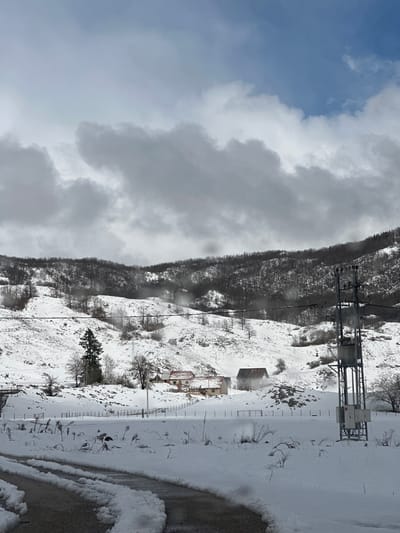 The image depicts a snowy winter landscape in Donja Brezna, Montenegro. The foreground features a partially snow-covered paved road with visible vehicle tire tracks. To the immediate sides of the road, undisturbed snow blankets the ground, dotted with sparse, dry vegetation. A black metal fence traverses the midground horizontally. Beyond the fence, two residential-style buildings are visible on a snow-covered rise: a light-colored structure with an orange-brown roof to the left, and a second, darker-roofed building to its right, from which white smoke or steam emanates. A tall, dark green coniferous tree stands prominently to the left of these structures. Power lines stretch across the midground, originating from a metal utility tower equipped with transformers and insulators positioned on the right side of the frame. The background consists of steep, snow-dusted hills heavily forested with bare deciduous trees, extending to the horizon under a heavily overcast sky dominated by large, dark grey clouds, with isolated patches of blue visible in the upper right. No individuals or animals are present in the scene.