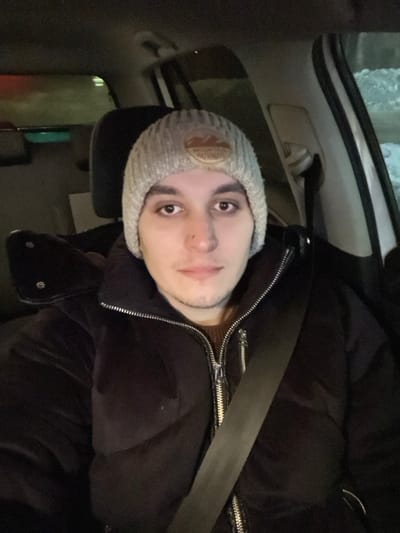 A young man wears a light gray beanie with a circular patch on it. He is wearing a dark puffy jacket with silver zippers, and a black seatbelt is visible across his chest. The interior of a car is visible behind him, including the car's roof, headrest, and the exterior through the window. He is facing the camera with a neutral expression.