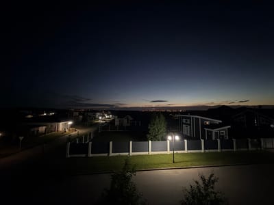 The image depicts a dimly lit suburban street scene in the village of Martemyanovo, Russia, during twilight or early night. Several modern houses with distinct architectural features are visible, characterized by dark siding and white trim. A dark fence with white pillars runs along the front of several properties, enclosing manicured lawns. Streetlights illuminate sections of the road and grassy areas. In the distance, a faint glow suggests a more populated area or city lights. The sky is a gradient of deep blue, transitioning to lighter hues near the horizon. No people are present in the frame.