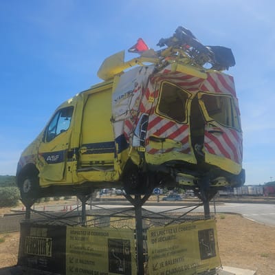 The image shows a severely damaged yellow vehicle, likely an emergency service or maintenance van, mounted on a metal platform outdoors. The vehicle is heavily deformed, with its roof and sides crushed and torn. Red and white reflective stripes are visible on the damaged sections. The vehicle is branded with "ASF" and appears to be a type of intervention vehicle. Two large yellow banners are attached to the fencing below the vehicle. One banner has text in French that translates to "WHEN WILL YOU HIT ME?" and a question mark. The other banner, also in French, reads "RESPECT THE SAFETY CORRIDOR." Below this, it instructs drivers to "When approaching an intervention vehicle: I slow down, I change lanes." The scene is set against a bright blue sky and a dry, scrubby landscape with some trees in the background. In the distance, there is a road and some parked trucks or shipping containers. The location is identified as Salles-d'Aude, France. The overall impression is a public awareness display about road safety and the dangers faced by intervention personnel.