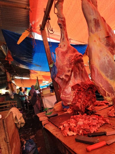 At a market, a butcher shop displays large cuts of meat hanging from hooks under a makeshift orange and blue tarp roof. On a wooden counter are piles of chopped meat and several knives. In the background, a few people are standing near the stall, including a woman wearing a hijab. The ground appears to be wet and cluttered with trash.