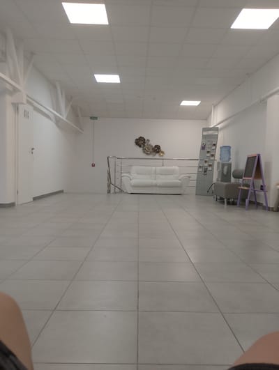 The image depicts an interior hallway or waiting area with a tiled floor and white walls and ceiling. A white sofa is visible in the middle ground, alongside a metal railing and stairs leading up. To the right, there is a water cooler, a brochure display stand, and a purple easel with a blackboard. The overall aesthetic is modern and minimalist. The scene is devoid of any people. The location is identified as Chaiky, Ukraine.