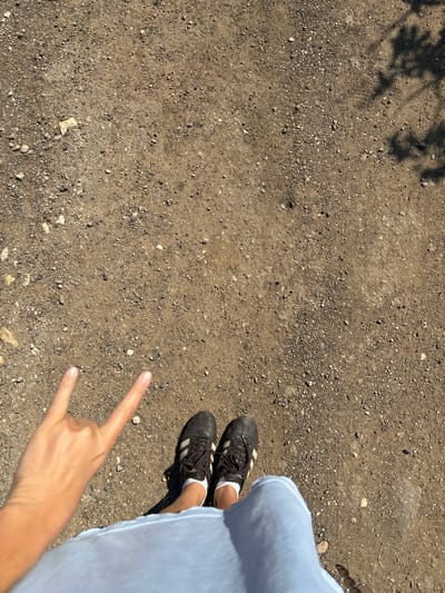 An overhead photograph captures a ground-level scene in Or Yehuda, Israel, featuring a partial view of an individual. The environment consists of dry, unpaved ground composed of light brown soil mixed with numerous small stones and gravel, under direct sunlight indicated by sharp shadows. In the lower center, a pair of black athletic shoes, featuring white soles and three distinct white stripes on the sides, are worn with white ankle socks. Brown laces are tied on both shoes. The shoes are positioned on the ground, with the toes pointing away from the camera. A light blue garment, likely shorts or a skirt, is visible partially covering the individual's lower legs and extending into the lower foreground. To the left of the shoes, a light-skinned hand is positioned, performing a "rock on" gesture with the index and pinky fingers extended upwards and the thumb, middle, and ring fingers curled. A dark, diffuse shadow, consistent with foliage or a tree branch, is present in the upper right quadrant of the frame.
