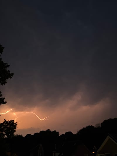 A vertical shot captures a dramatic lightning strike illuminating the clouds and silhouetted trees and rooftops in Putten, Netherlands. The sky is a dark, stormy grey, but the lightning branches out in vibrant yellow and orange hues across the mid-section of the frame. The lower part of the image shows the dark silhouettes of trees and the peaked roofs of houses, with one house in the foreground having a distinctly pointed roof and a visible light within. The overall atmosphere is dark and ominous, with the powerful natural phenomenon taking center stage.