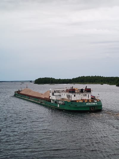 This media file depicts a barge, identified by the visible text "НЕВСКИЙ-6", transporting a large quantity of sand. The barge is green with a white superstructure. The scene is a wide river or body of water, with a dense forest lining the distant shore. Several other smaller vessels and distant landforms are visible in the background, suggesting a navigable waterway. The sky is overcast with a light, diffused light, indicating either early morning, late afternoon, or a generally cloudy day. The water is calm with gentle ripples. The overall atmosphere is peaceful and utilitarian, focusing on the transport of cargo. The location is identified as Novokemskii, Russia.