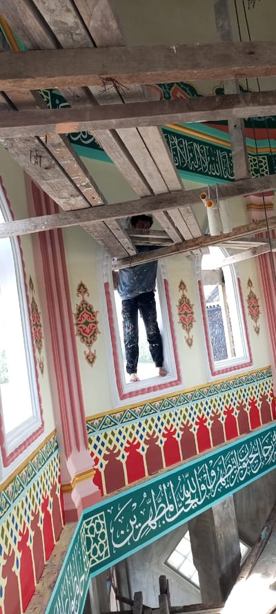 This image depicts the interior of a building under renovation in Kumbang, Indonesia. A barefoot male worker, wearing a paint-splattered blue long-sleeved shirt and black pants, stands on the sill of a rectangular window. His upper body is visible above a horizontal wooden scaffolding beam, with his head turned towards the viewer.

The cream-colored walls are extensively decorated with multi-colored Islamic patterns and calligraphy. A prominent teal green horizontal band features white Arabic calligraphy. Below the windows, a patterned band displays red, maroon, and blue arch-like motifs interspersed with multi-colored diamond designs (gold, green, blue). Above the windows, intricate red, green, and gold floral patterns are painted. Multiple rectangular windows with white frames and pink sills admit natural light, revealing a muted green exterior and part of an adjacent roof structure. Pink fluted pilasters separate these windows.

The upper portion of the scene is dominated by a dense network of rough-hewn wooden beams forming temporary scaffolding, some with exposed nails. A white PVC pipe with two yellow end caps is suspended from one of these beams. The ceiling, partially visible behind the scaffolding, shows light green and patterned sections.