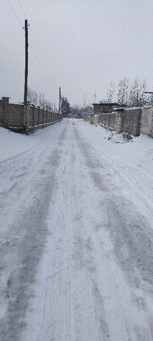 A snow-covered road stretches toward the horizon, marked with tire tracks and patches of ice. A concrete barrier lines the left side of the road, with leafless trees visible beyond it. An electrical pole stands near the barrier, wires extending from it. The scene is dominated by gray and white tones, creating a bleak, wintry atmosphere. The image orientation is sideways.