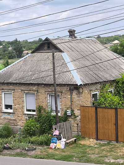 The image depicts a residential property in Yasynivka, Ukraine. A brick house with a corrugated metal roof dominates the foreground, showing signs of age and weathering. Several power lines crisscross the sky above the house. Adjacent to the house is a wooden fence and a collection of various items, including backpacks, bags, and buckets, placed on the ground near a wooden bench. The yard is overgrown with vegetation, and the scene appears to be a typical residential setting in a rural or semi-rural area.