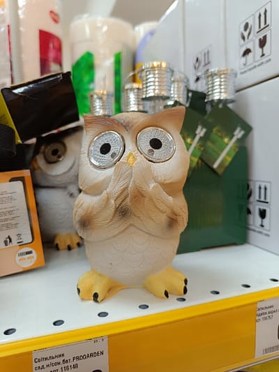The image shows a white owl figurine with large black eyes and a yellow beak. It is sitting on a yellow shelf in a store, with the owl's head tilted to the side. There are two boxes with product information on the shelves. The boxes and the owl are on a shelf in a store with several other items visible in the background. The store is located in the city of Berehove, Ukraine, at the coordinates 48.194, 22.655. The owl is likely a garden ornament.