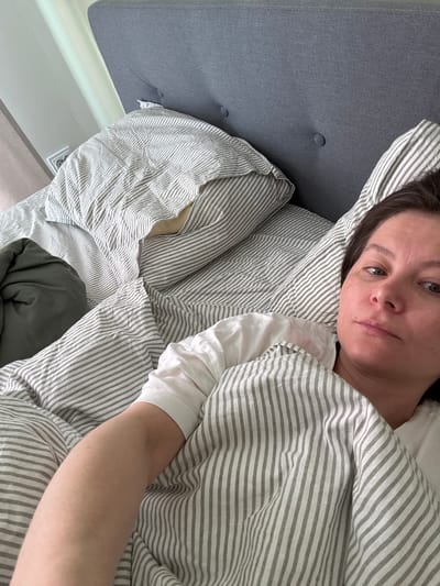 A woman lies in a bed with striped bedding. Her head rests on a pillow with similar striped fabric. She is partially covered by the bedding and appears to be wearing a white top. Her arm lies extended on top of the bedding. A gray headboard is visible on the left side of the image. The location in the city of Люк, Russia is not discernible from the image.