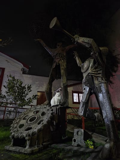 A white cat sits on a large, weathered metal sculpture in an outdoor setting at night. The sculpture appears to be made of recycled industrial metal and depicts two humanoid figures with abstract, angular forms. One figure on the left holds a large, spherical object, while the other on the right appears to be holding a horn or pipe. A large, toothed metal wheel, possibly part of a machine or vehicle, is positioned in the foreground, serving as a base for the sculpture. The cat is the only living subject in the frame. Behind the sculpture, a white building with red trim and windows is visible. The ground is covered in grass and moss. The lighting suggests artificial illumination, creating shadows and highlighting the textures of the metal. This scene is located in the city of Chillán, Chile.