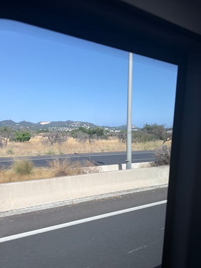 The image shows a view from a vehicle, looking out of a window. In the foreground, a dark grey asphalt road with a white line marking the lane is visible. To the left of the road, a concrete barrier separates it from a dry, grassy area with sparse vegetation. A tall, slender grey pole stands vertically on the other side of the barrier, partially obscuring the view of the background.
The background features a landscape of rolling hills with scattered buildings, suggesting a semi-rural or suburban environment. The hills are covered in dry, yellow grass and some scattered trees. The sky above is a clear, bright blue. The overall impression is that of a sunny day, possibly in a Mediterranean climate, given the dry vegetation and the architecture visible on the hills. The image appears to be taken while the vehicle is in motion, as indicated by the slight blur in the foreground. The specific location of Santa Eulària des Riu, Spain, is not explicitly identifiable from the visual elements alone.