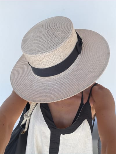 A person wearing a wide-brimmed straw hat with a black ribbon is shown from the chest up, against a plain white background. The hat obscures their face. They are wearing a black and white striped dress or top. A black bag is slung over one shoulder, with a thick rope strap visible. The setting appears to be outdoors, possibly a patio or balcony. There is no clear indication of the location being Baan Bo Phut, Thailand.