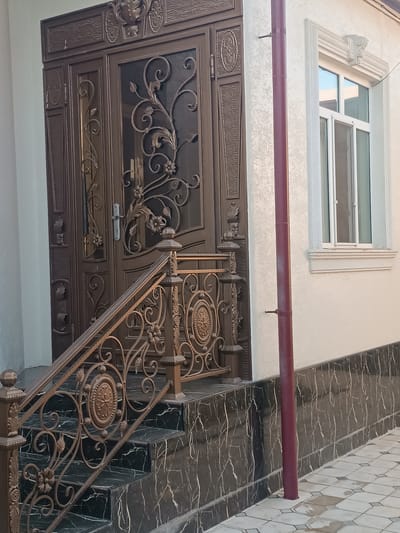 This image shows a close-up, eye-level view of a portion of an entryway in Shamsiabad, Uzbekistan. The central focus is a large, ornate double door made of dark bronze metal. The doors feature intricate scrollwork, floral motifs, and decorative panels. A silver door handle is visible on the left door. To the left of the door, a dark bronze metal staircase with a matching railing leads up to the entrance. The steps of the staircase are covered in dark, polished stone with white veining.
To the right of the door and staircase is the exterior wall of a building. The wall is painted a light, textured color. A burgundy-colored downspout runs vertically down the wall, attached by a small bracket. Above the downspout and to the right of the door is a white-framed window with multiple panes. The window is topped by a decorative stone molding. A beige stone ledge runs below the window.
The lower part of the building's wall is clad in the same dark, polished stone with white veining as the staircase. In the foreground, a paved walkway with light-colored square tiles, some of which appear wet, extends from the bottom of the frame. The overall impression is of a well-maintained and elaborately decorated residential entrance. There are no people or animals visible in the image.