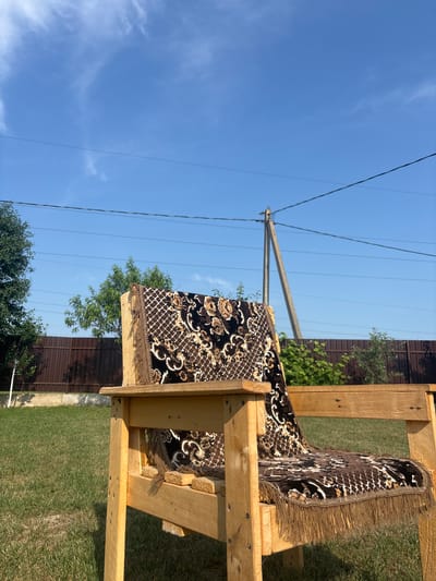 This image depicts an outdoor scene in Skidzieĺ, Belarus, featuring a wooden chair with a patterned textile draped over it, placed on a grassy lawn. The chair is constructed from rough-hewn wooden planks. The textile, which covers the backrest and seat of the chair, has a brown and white intricate pattern with fringed edges. In the background, a brown wooden fence runs horizontally, with green foliage visible behind and in front of it. A utility pole with power lines stretches vertically in the mid-ground. The sky above is blue with scattered wispy white clouds. The overall setting suggests a rural or suburban backyard.