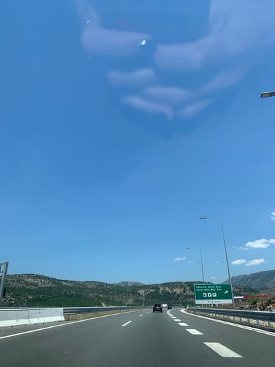 The image captures a view from a highway in Gornje Mrke, Montenegro. A black car is visible driving on the highway in the distance. To the right, a green highway sign indicates "Odmorište Gornje Mrke - Gornje Mrke Rest Area" with icons for gas, food, and restrooms, and an arrow pointing right, with "NEXT 45 km" indicated. The highway is bordered by a metal guardrail and a concrete barrier. In the background, a mountainous landscape covered in trees and shrubs is visible. The sky is clear blue with some light clouds.
