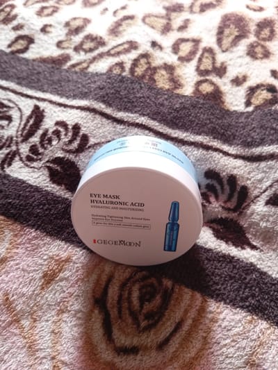 A white container of eye mask with hyaluronic acid is placed on a tan and brown leopard-print blanket. The container has text in English and Russian. The English text reads: "EYE MASK HYALURONIC ACID HYDRATING AND MOISTURIZING Hydrating Tightening Skin Around Eyes Improve Eye Dryness It gives the skin a soft, smooth radiant glow GEGEMOON". There is also a small image of a blue vial on the container. The background is a blurry leopard-print blanket. The image is taken from a top-down perspective. There are no people or animals visible in the image. The location of Monastirische, Russia is not depicted in this image.
