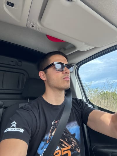 A man with short dark hair and a trimmed beard is wearing black sunglasses and a black T-shirt with a design on the front and a logo on the sleeve that reads "KKK CLUB KIRKOVO." He is wearing a seatbelt and appears to be in a vehicle with an open sunroof, as the sky and some trees are visible above. The interior of the vehicle is light gray. The photo suggests the location is in or near Kirkovo, Bulgaria due to the "CLUB KIRKOVO" logo.