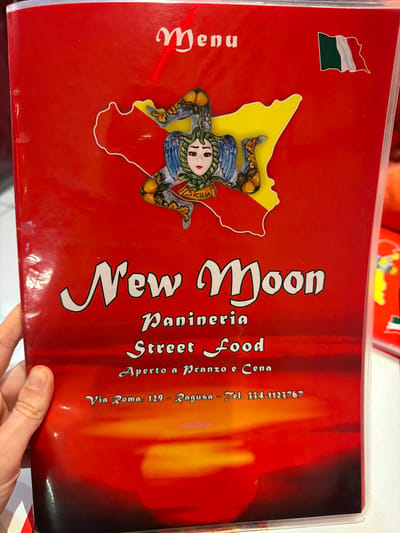The image shows the menu of a restaurant called "New Moon Panineria Street Food" located at Via Roma, 129 in Ragusa, Italy. The menu is red with the name "New Moon" in large white letters. The restaurant offers street food and is open for lunch and dinner. The menu also features a symbol of Sicily, with the "Trinacria" in yellow outline on a red map background, as well as a Hungarian flag in the upper left corner. The telephone number is 334.1123767. A hand is holding the menu.
