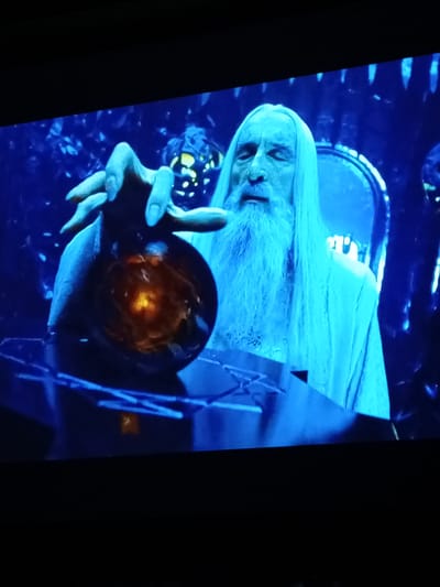 The image shows a close-up of a man with a long white beard and pale skin, wearing a white robe. He has his eyes closed and is holding a dark orb in his left hand, which contains a glowing orange light. The background is dark and appears to be a dimly lit interior with blue lighting. The man is seated, and part of a throne or ornate chair is visible behind him. A platform or table is in front of him, on which the orb appears to be resting. The scene is dark with a blue cast, suggesting it is a dramatic or mystical moment. The specific location of Zam'yany, Russia, is not depicted in this image.