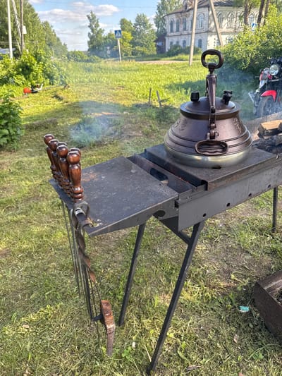 An outdoor scene in Луптюг, Russia, captures a cooking setup under bright daylight. In the foreground, a large, dark-colored, ornate pot with a distinctive handled lid sits atop a black metal brazier or grill stand. Smoke gently rises from beneath the pot, indicating a fire is burning and cooking is in progress. The pot appears to be made of cast iron or a similar heavy material, featuring embossed decorative patterns around its body.
To the right of the pot, an array of metal skewers with polished wooden handles is neatly arranged on a rack, suggesting preparations for grilling alongside the pot-cooked dish. The brazier itself is a sturdy black metal structure with several vents or holes along its sides.
The setting is a grassy area. In the middle ground to the left, a large, weathered multi-story building with multiple arched windows and intricate architectural details stands, hinting at a historical or traditional local structure. Further back, a red and black motorcycle is partially visible, parked on the grass. The background is dominated by lush green trees and foliage under a blue sky with scattered white clouds. A blue road sign with a white arrow is visible in the distance, suggesting proximity to a road. The overall impression is a casual outdoor cooking event, possibly a picnic or a gathering in a rural or semi-urban environment. No people or legible text are visible.