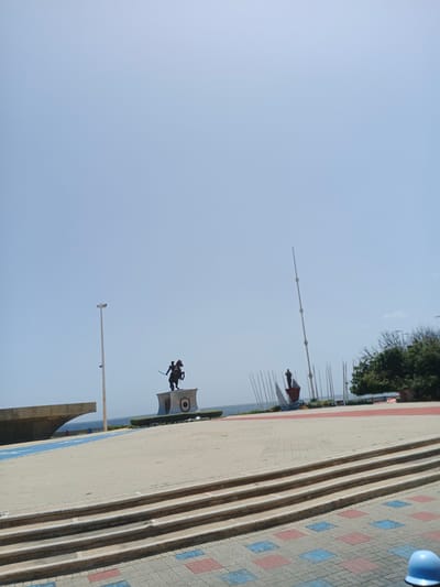 The image depicts a wide paved plaza with a clear blue sky overhead. In the middle ground, a bronze equestrian statue of a man on horseback stands atop a pedestal. To the right of the equestrian statue, a smaller figure stands on a boat-like structure. Several tall, thin poles are arranged in the background, extending into the sky. In the foreground, there are tiered steps leading up to the plaza. The ground is paved with rectangular bricks in red, blue, and gray patterns. A light blue hard hat rests on the lower right corner of the image. The background shows a body of water, likely the sea, and some trees and structures, suggesting an outdoor urban setting. This location is the Plaza Mayor in La Guaira, Venezuela, a prominent public space featuring historical monuments and overlooking the Caribbean Sea.